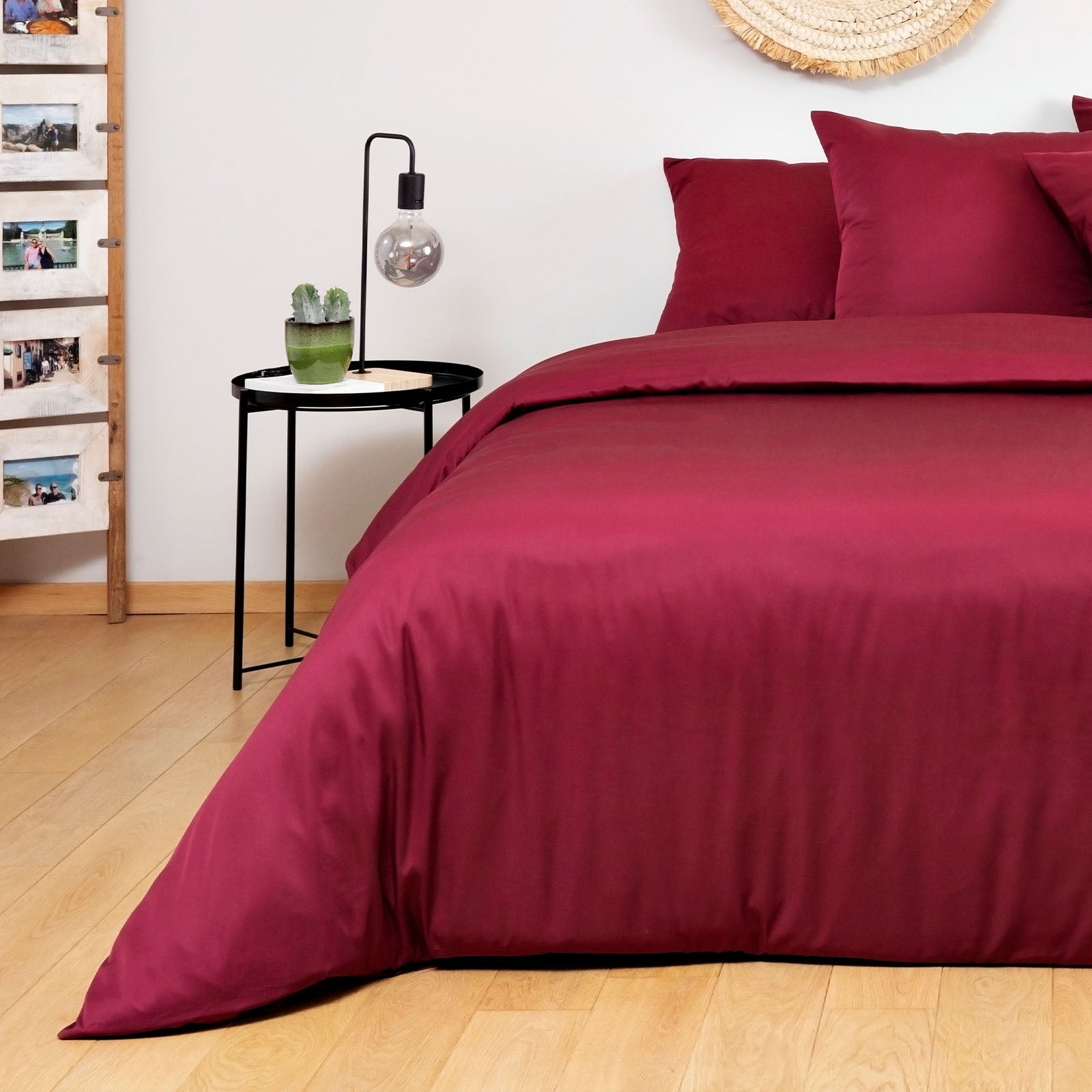Burgundy bedding set on a bed in a room with wooden flooring and a side table.