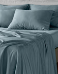A neatly made bed with dusty blue bambaw cozy bamboo bedding, including a fitted sheet, pillowcases, and a matching duvet, styled with several pillows against a light neutral headboard.