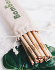 Natural bamboo drinking straws in a drawstring bag placed on a marble surface with green leaves