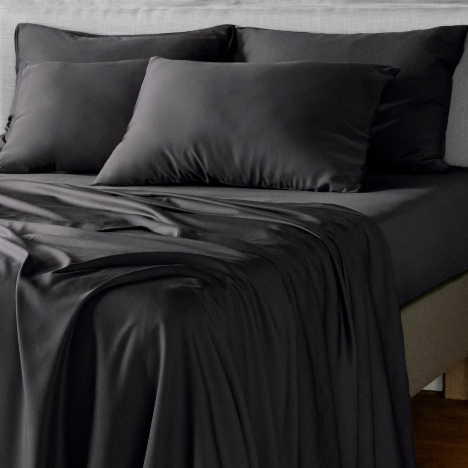 Black bamboo bed sheet and pillowcase set on a bed with rumpled fabric, shown in a dark studio setting.
